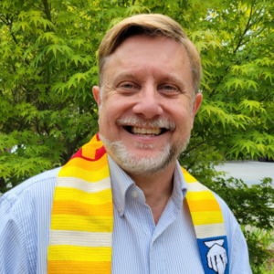 Rev. Michael Cronin, white male with blonde hair and a grey beard, wearing a light blue and white vertically striped shirt with a yellow stole around his shoulders